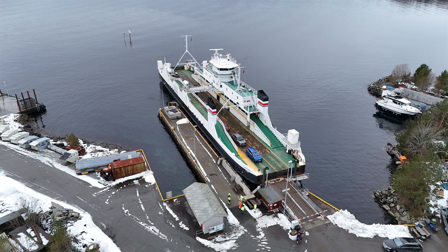 Vi ser ei ferjekai frå lufta med land i forgrunn og grå sjø bak. Ein smal veg går ned til kaia, der ligg det ei ferje inn til ei smal utstikkande kai. Ferjebrua er og smal og lita, og mykje mindre enn det ferja eigentleg treng i høve si breidde. Ferja er lita, kvit med grønt dekke, med sidestilt styrehus. Bommen er nede og 5 bilar er synlege på ferja. det er snø på bakken og mykje rust på kai og ferje. 