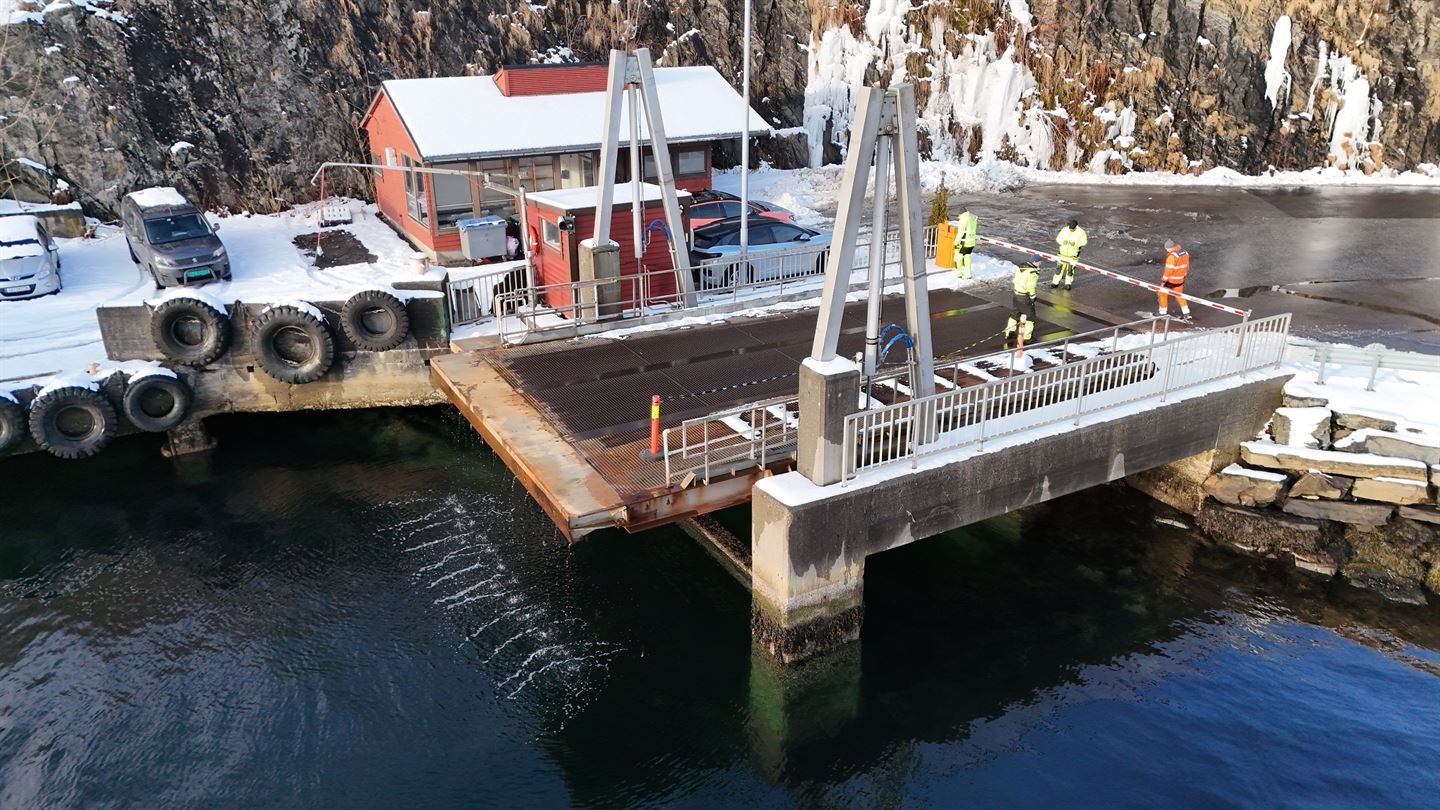 Bilde frå lufta (drone) av ei lita ferjekai. blå sjø i forgrunnen og parkeringsplass med to bilar i bakgrunnen og asfalterte biloppsitllingsfelt og veg inn mot kaia bak til høgre. Eit lite raudt trehus er rett bak kaia, venterom. Bak dette er det fjellskjæring, og landskapet er og dekka av snø. Dagslys og sol. Tre personar i gult arbeidstøy og ein i oransje arbeidstøy går p åkaia og inspiserer. Kaia har tydeleg og mykje rust. 