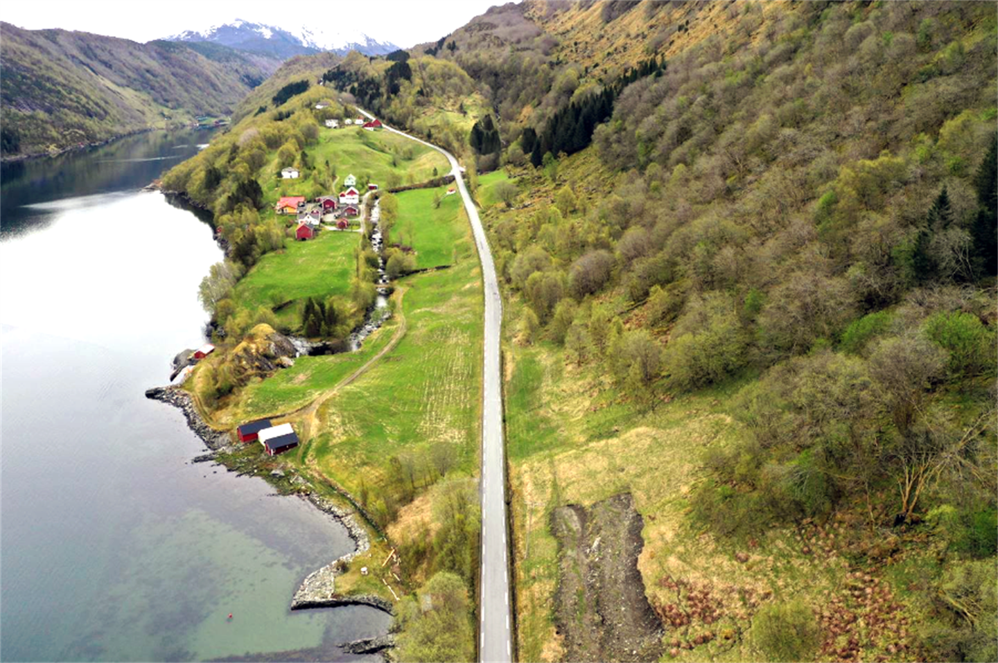 landskapsbilde sett frå lufta, ein blå fjord går innover landskapet, ein smal asfaltert veg går langs fjorden og over eit berg mellom fjorden og ei bratt skogkled fjellside. Grøne bøar og nokre husklynger og fjøs langs vegen. 