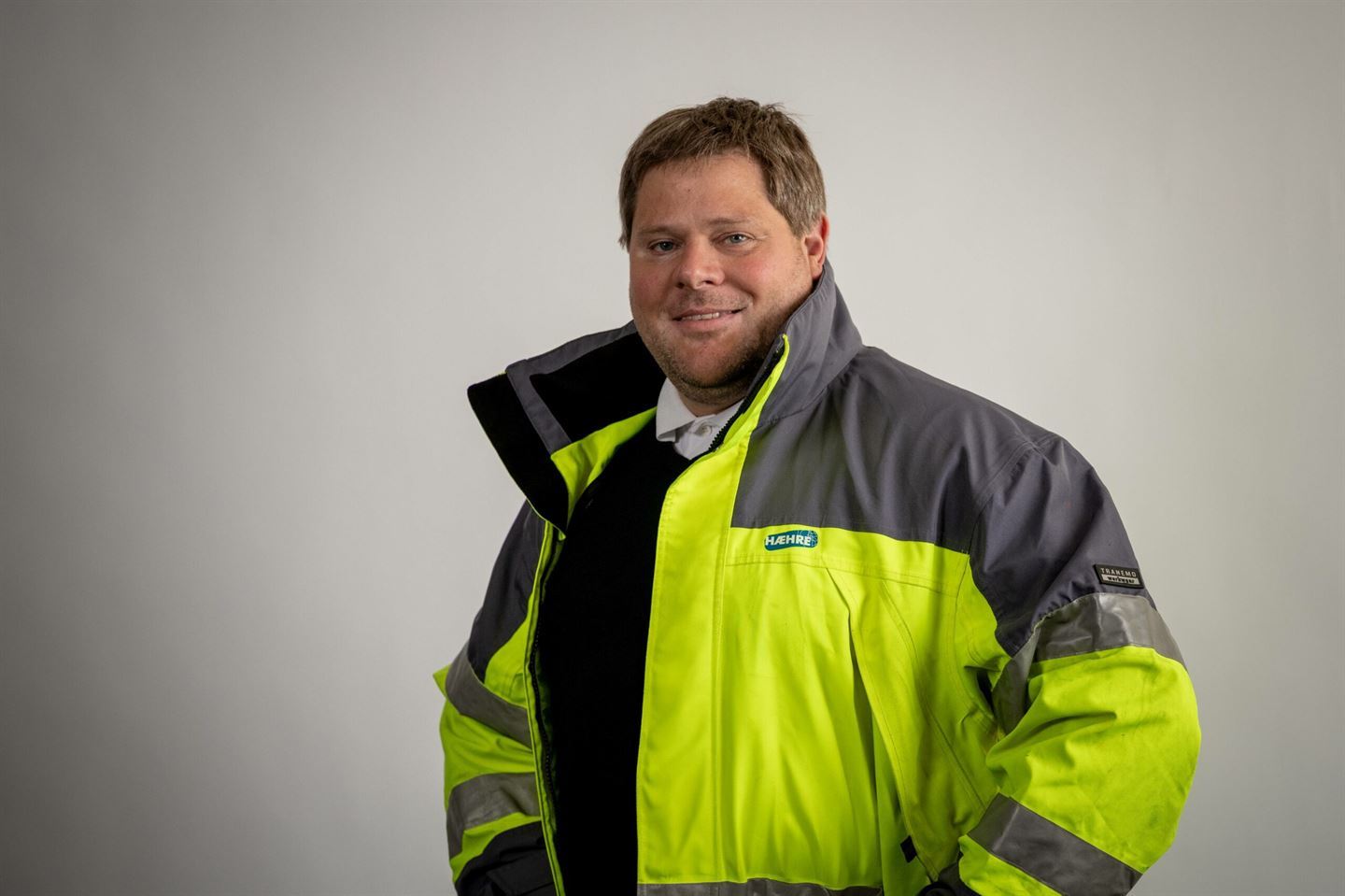 Vi ser eit profesjonelt tatt portrettbilde av ein mann frå livet og opp, ikledd ytterjakke i svart og signalgult - ei typisk arbeidsjakke for vegarbeid. Han har kort blondt hår og smiler mot kamera. Nøytral bakgrunn. 