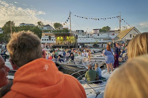 Hamneområde, der mange folk og båtar er samla. I bakgrunnen er det ei scene med lys og artistar.