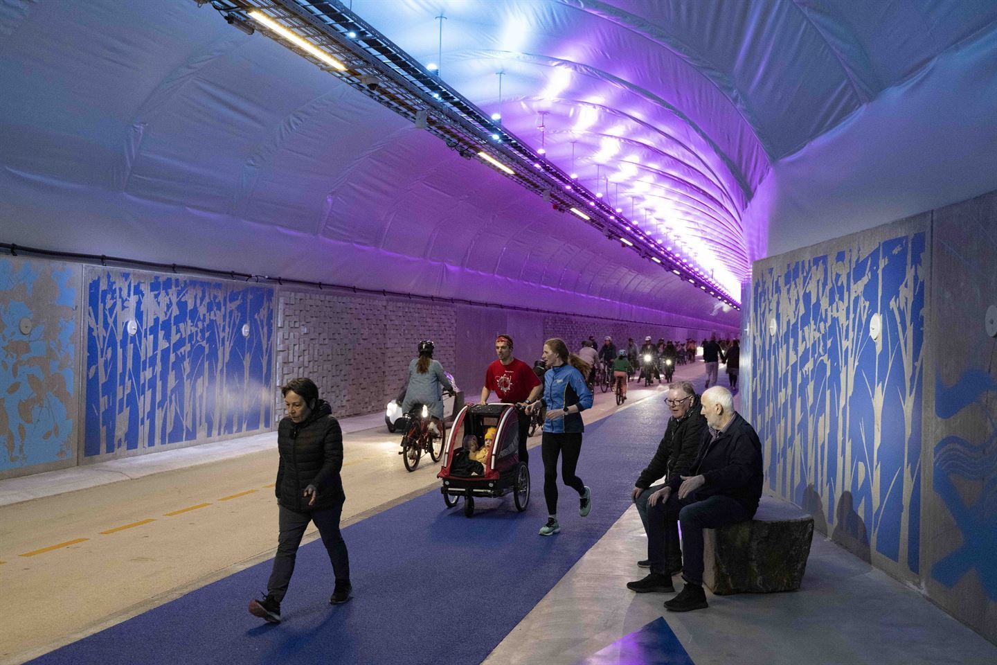 Bilde fra inne i ein tunell. Tunellen er for gåande og syklande, med grått dekke på ein halvdel av vegdekket og blått dekke for gåande på andre halvdel. I forgrunnen ser vi ei eldre dame i svart boblejakke gå framover, fulgt av ei yngre par mann og dame joggande bak skyvande på ei sykkelvogn med to born. På høgre side av tunellen sit det to eldre menn på ein benk. Veggane i tunellen er dekka av heildekkande kunstverk i grått og blått og turkis og grå steinmur. Taket er heildekka av eit plastdekke med gjennomgåande lys som nokon stader kastar lilla lys i taket. Vi ser fleire gåande og syklande i tunellen der den strekk seg bakover i bildet. 