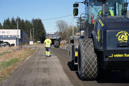 Ein mann i gule arbeidsklede går midt på ein grå grusveg. Bak han til høgre i bildet er ein stor svart anleggsmaskin med gul skrift. Foran mannen er ein annan stor anleggsmaskin. 