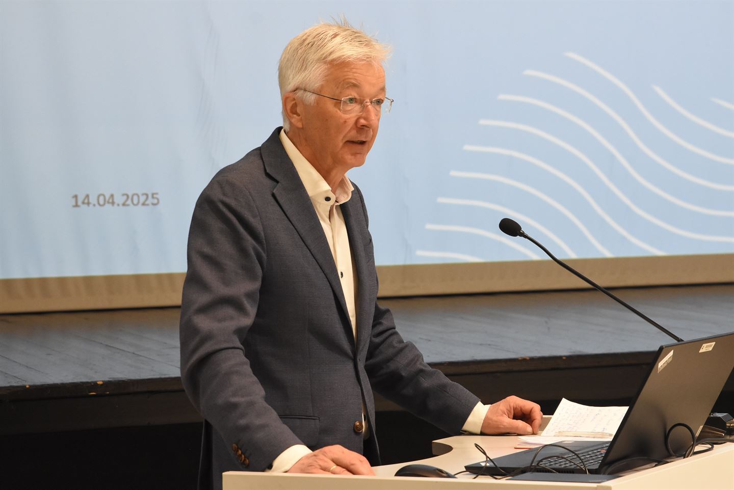 Ein mann med grå dress og lyst grått hår står på ein talarstol og se utover ein forsamling. 