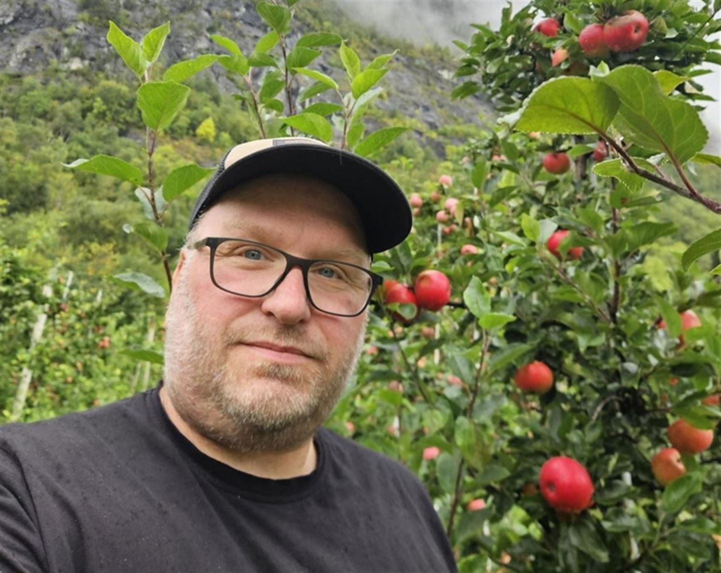 Ein mannn med briller og svart caps og svart tskjorte står foran ein hage med epletre. Bak han er eit fjell og grå himmel. Foto: privat