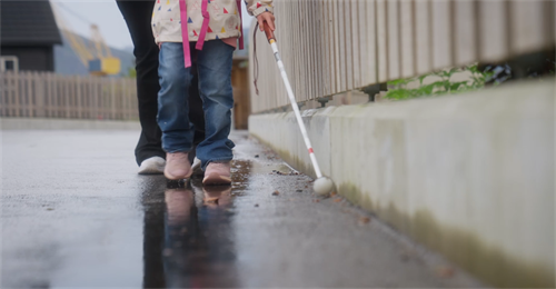 To par bein går på fortauet langs ein veg. Foran går eit barn med lyserosa joggesko, blå dongeribukse og kvit jakke med fargerike trekantar på. I handa held ho ei kvit blindestokk. 