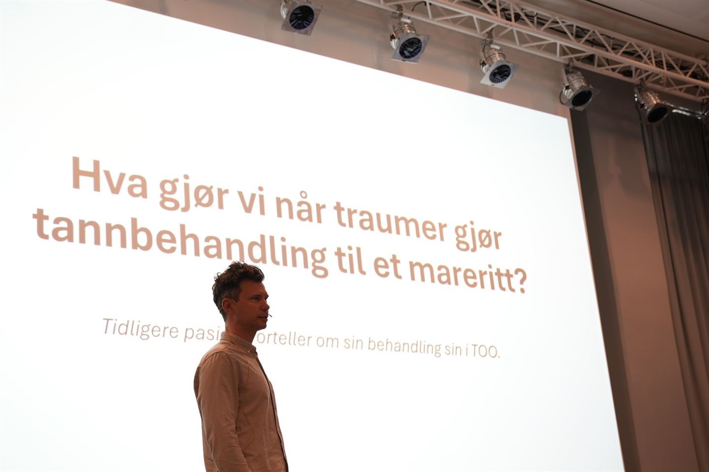 Psykolog John tveit-winther står i en konferansesal, foran en storskjerm der det står "hva gjør vi når traumer gjør tannbehandling til eit mareritt?"