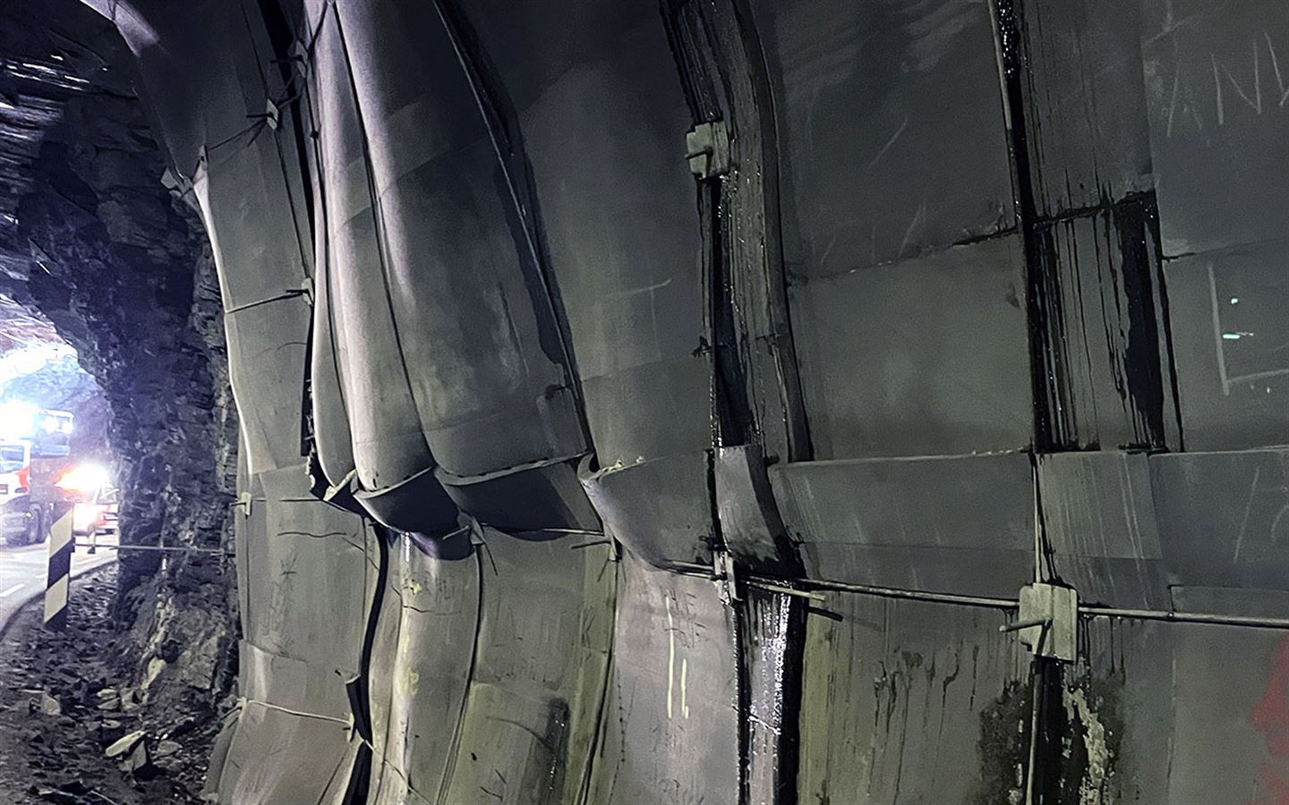 Ein tunellvegg er dekka av dårleg festa grå plater festa med boltar, over ein grå smal dårleg asfaltert veg. Vi ser så vidt utgangen av tunellen og ein bil som står utanfor til venstre i bildet. Over platene er det grovt hakka fjellvegg. Fleire plater bular ut, noko bak dei sprenger seg ut. 