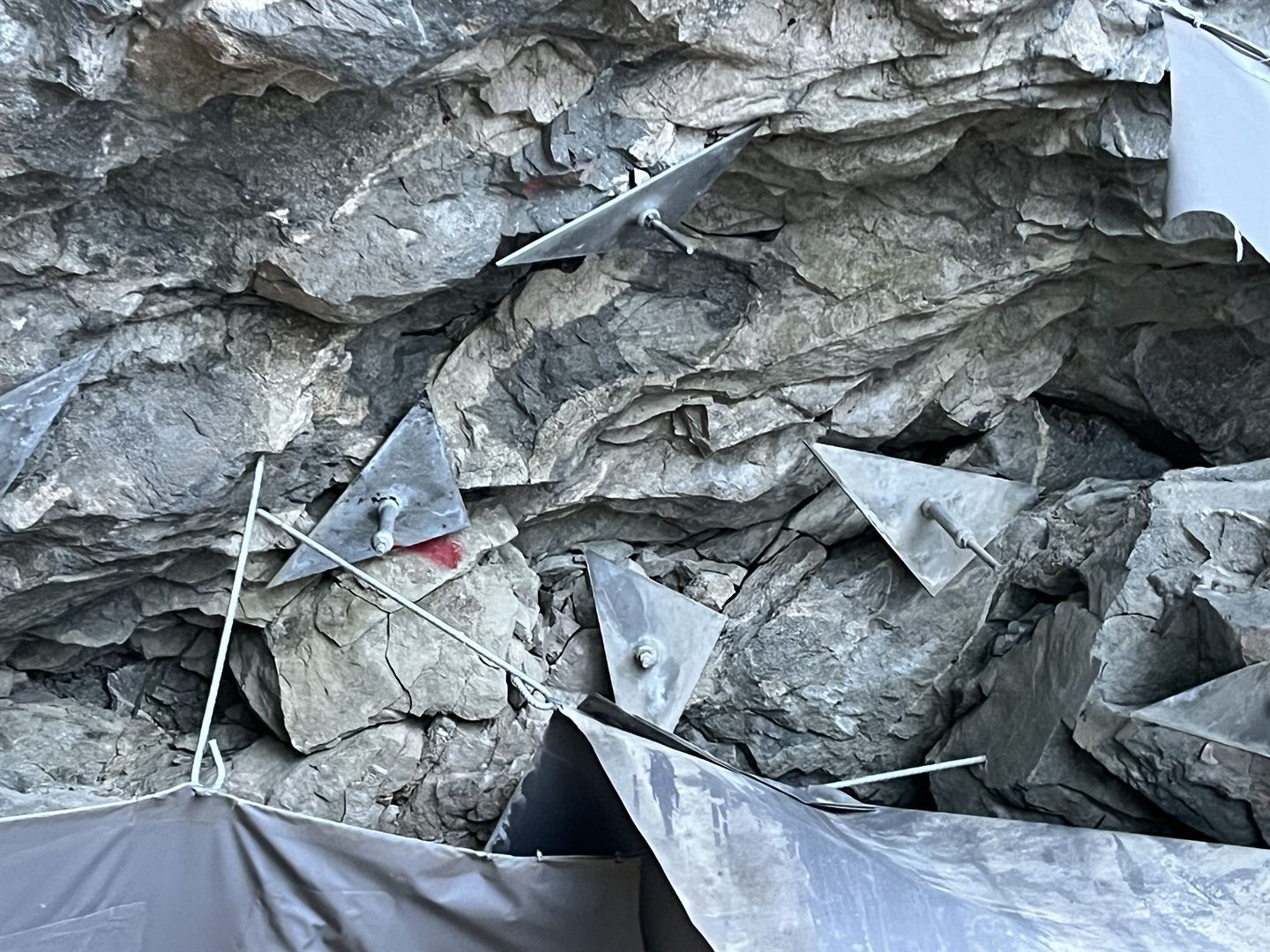 Vi ser del av taket i ein steintunell. Fjellet er grovhogd og det ser ut som det kan rase ut delar og steinar av fjellet når som helst. Vi ser fleire boltar som er bora inn i fjellet, men som no er lause, og restar av ein duk. 