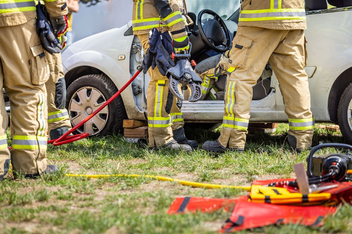 Brannkonstablar i ei trafikkulykke. I bakgrunnen er eit bilvrak. Alle konstablane er avbilda med berre beina. Det ligg ulike verktøy foran i bildet. 