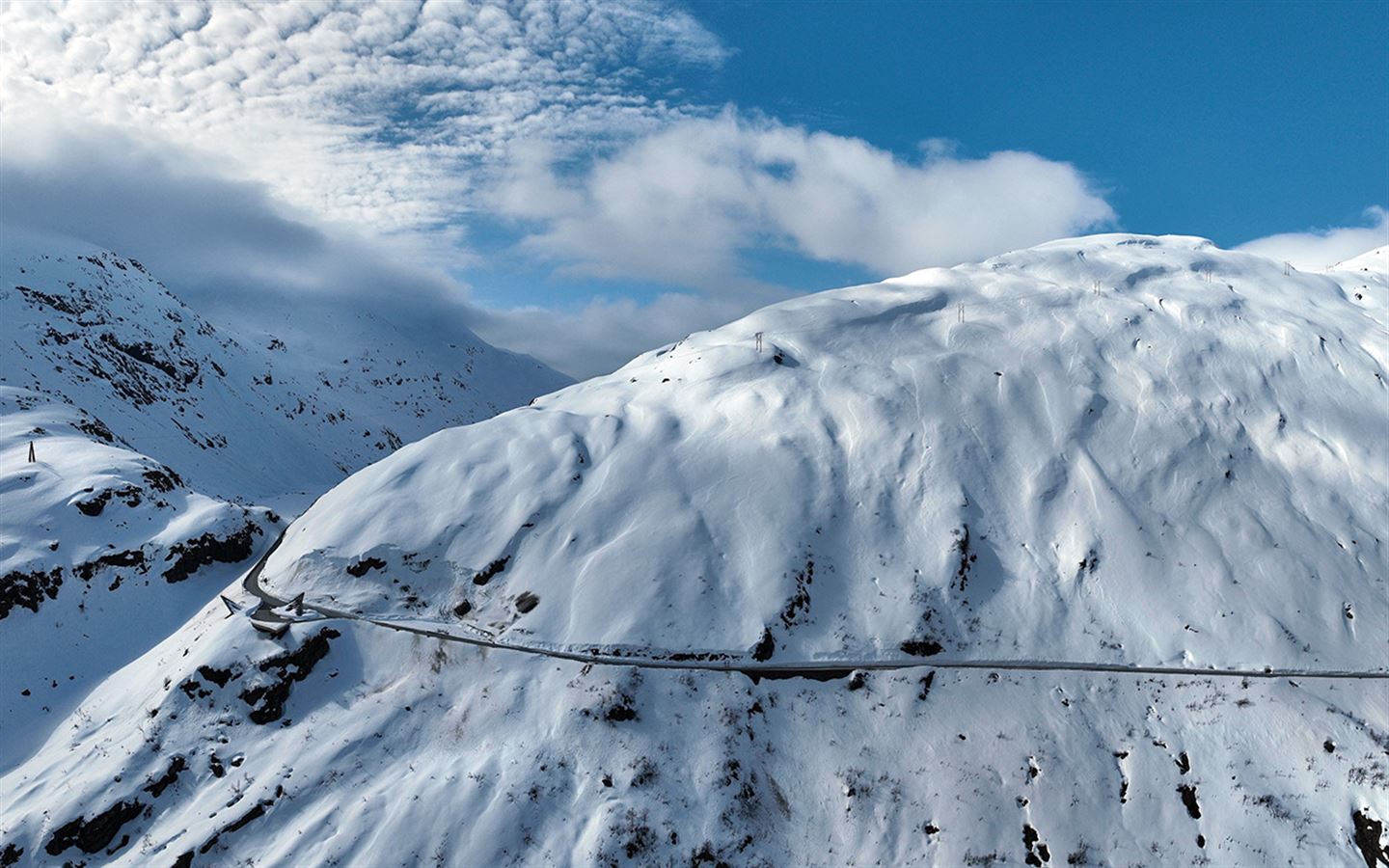 Bilde frå oven/lufta av ei snødekt fjell, blå himmel bak. Ein svart veg skjer gjennom framsida av fjellet og snur inn i fjellheimen på venstre side, i svingen er det eit flott bygg og ein parkeringsplass med utsiktspunkt. 