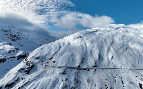 Bilde frå oven/lufta av ei snødekt fjell, blå himmel bak. Ein svart veg skjer gjennom framsida av fjellet og snur inn i fjellheimen på venstre side, i svingen er det eit flott bygg og ein parkeringsplass med utsiktspunkt. 