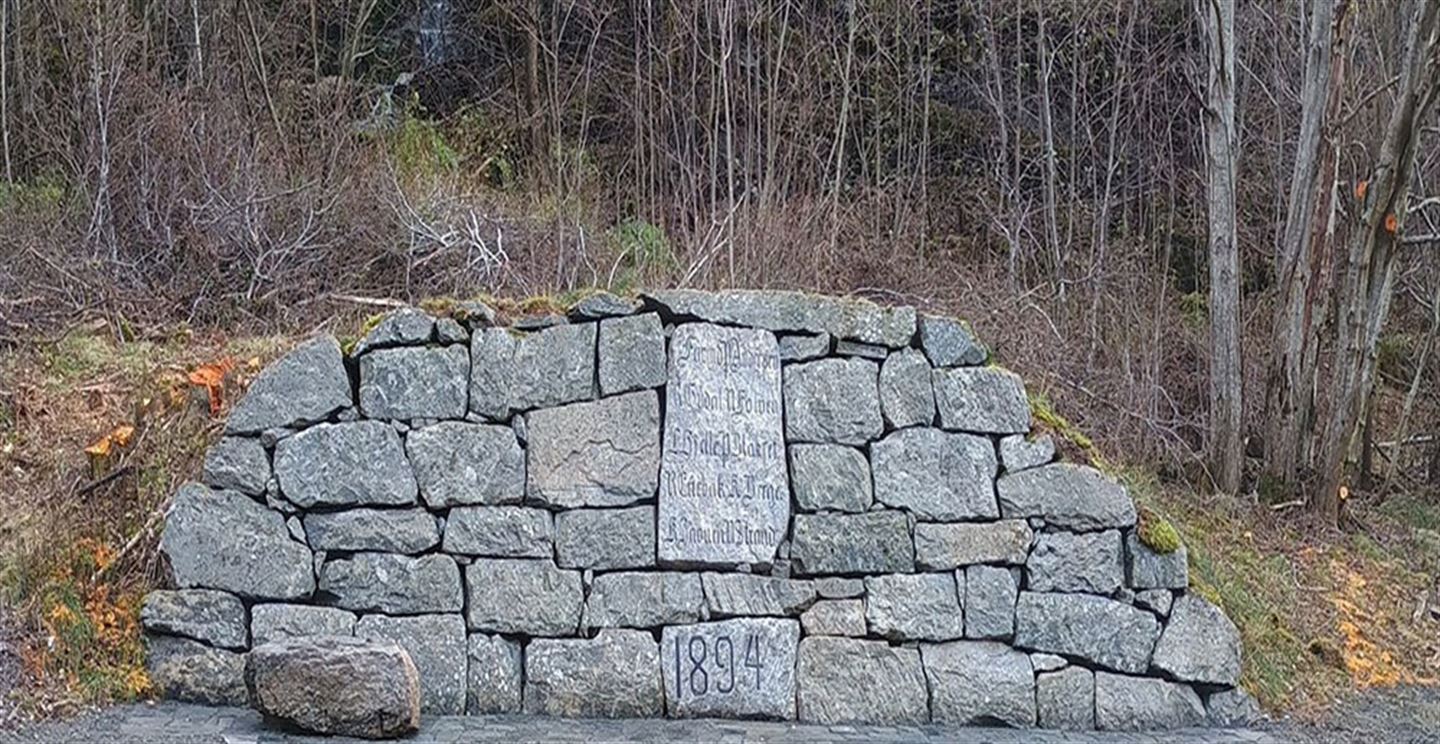 Steinmur med inskripsjon i stein, heller framfor og ei vassblokk ståande på hellene. 