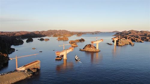 Landskapsbilde der vi ser eit kystlandskap frå sjøsida, med fleire holmar og skjer i fint gyllent solnedgansglys. Det blir bygd ei bru i landskapet, vi ser enden på ein anleggsveg til venstre i bildet, og tre store begongsøyler i havet og på ein holme der brudekket blir støypt meter for meter. Det er ei oransje heisekran ved kvar søyle. Søylene er omlag 50 meter høge. blå himmel og blått hav, brune holmar utan tre eller vegetasjon. 