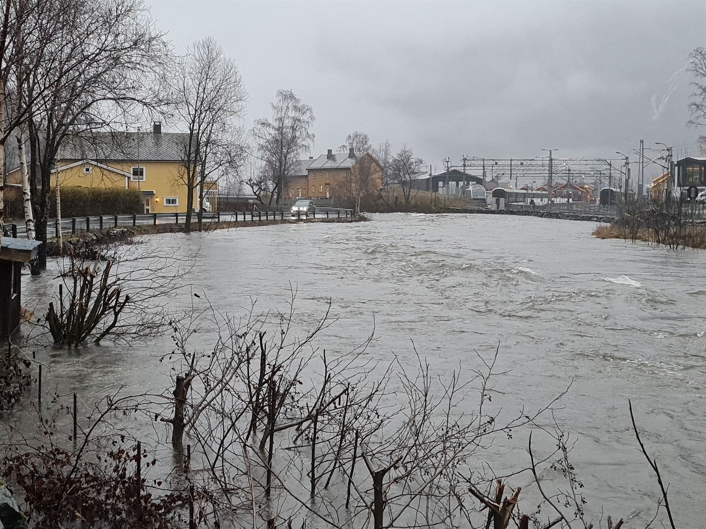 Bilde av ei brei elv der vatnet går nesten heilt opp til vegen. Trer, buskar og gjerde står i vatn. Bak i bilde ligg ein jernbanestasjon. 