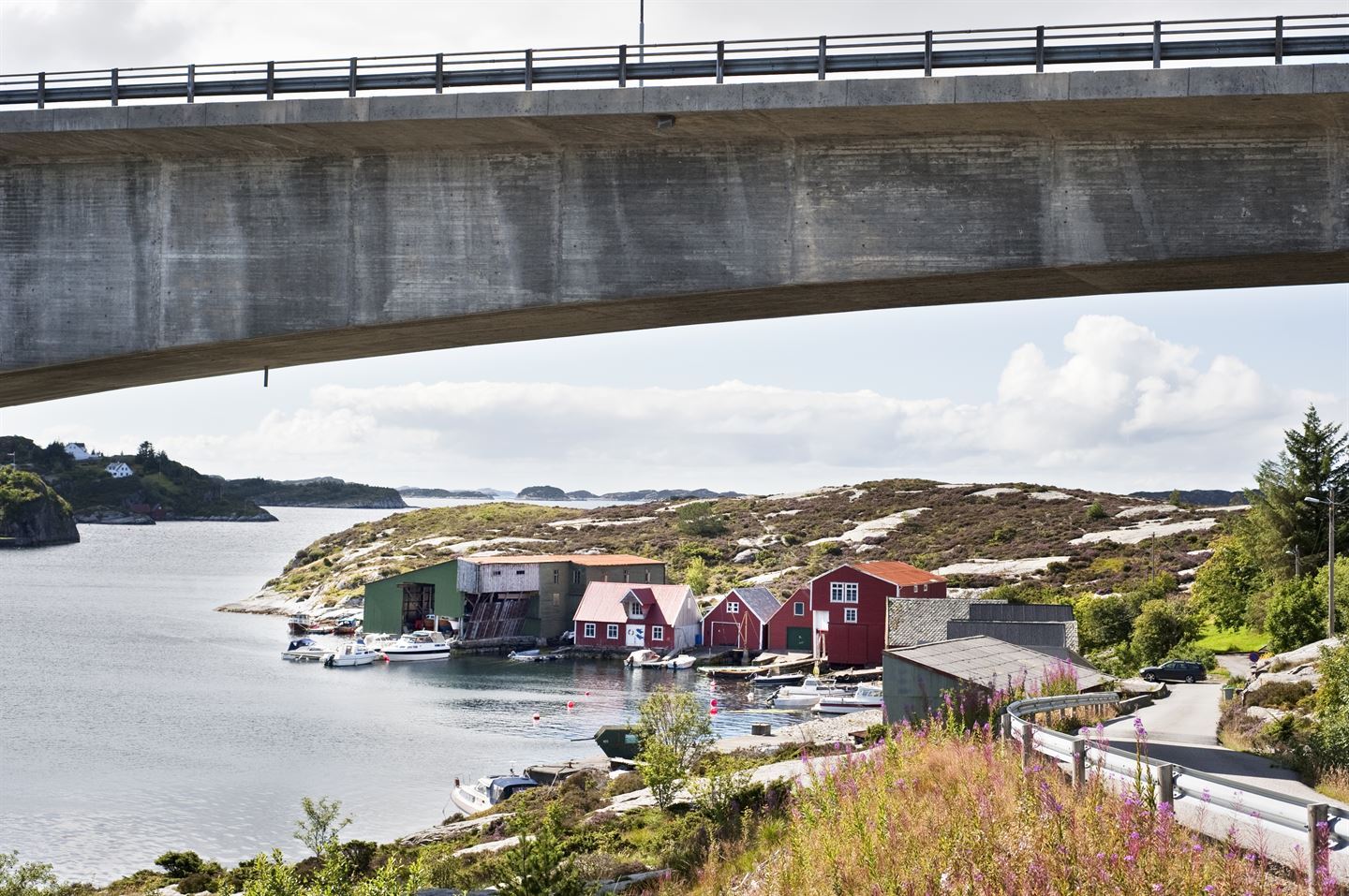 Ei grå bru går på tvers i toppen av bildet. I bakgrunnen, under brua, ligg det grøne og raude naust ved sjøen. Foran dei ligg det små båtar. Ein smal veg går under brua.