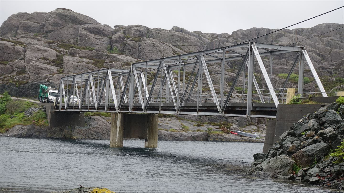 Ei grå stålbru med bjelker. Bak brua er det berg og under brua er det vatn. 