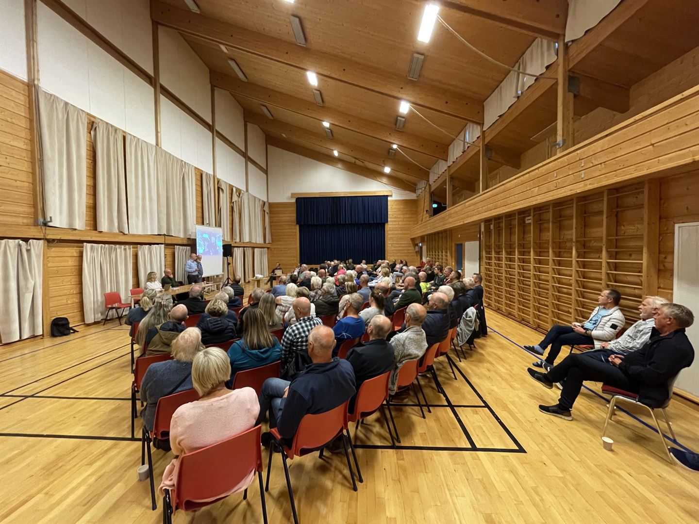 Idrettshall med parkettgolv og trepanel på veggane. Salen er avlang og i eine enden enden er det ei scene. På langs av rommet er det sett opp stolar tett i tett, og på langsida er det eit lerret og ein prosjektor. Det sit menneske på stolane, og på sida av lerrretet er det eit bord. Ved bordet sit det ein mann og ei kvinne, to menn står ved sidan av lerretet.