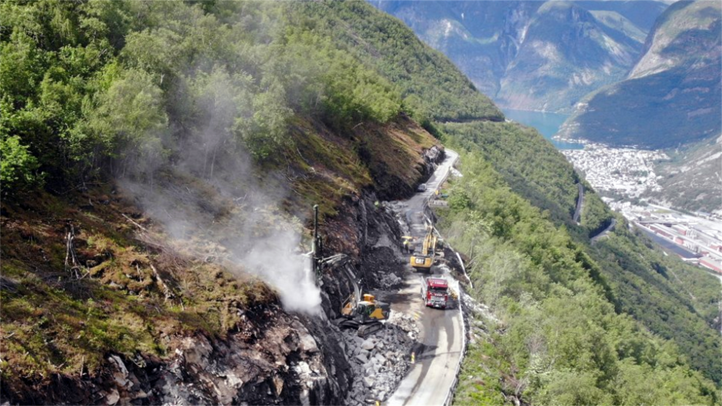 Luftbilde av vegarbeid i bratt fjellskråning på fylkesveg 53 med tettstaden Årdal i bakgrunnen. Gravemaskin og lastebil er i arbeid på vegen, og det kjem røyk frå inngangen til det som skal bli ein tunnel, Ljoteli-tunnelen. Det er sprengt ein del i fjellsida før tunnelen. Vår, grøn fjellside med skog og blå himmel. 