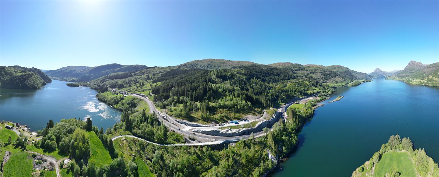 Dronefoto som viser fjord, med veg som slunger seg langs fjorden og fjell bak. Det er sommar, sol og grønne lier.