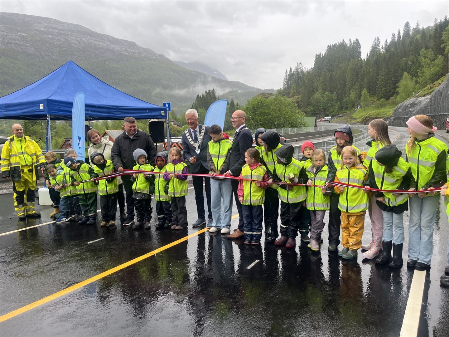 Opning av veg med gul midtstripe ein regntung dag i mai. Mann med fylkesordførarkjede held i snora, med mange ungar i regnkle og gule refleksvestar står rundt han og held i snora.