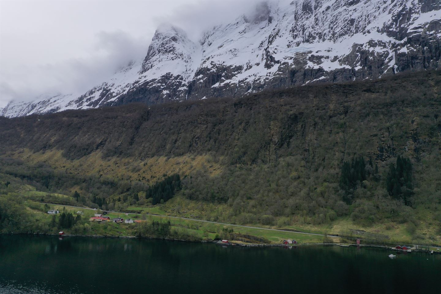 Haustmotiv. Ein veg og hus som ligg ved fjorden, med bratt fjellside og snøkledde fjelltoppar bak.