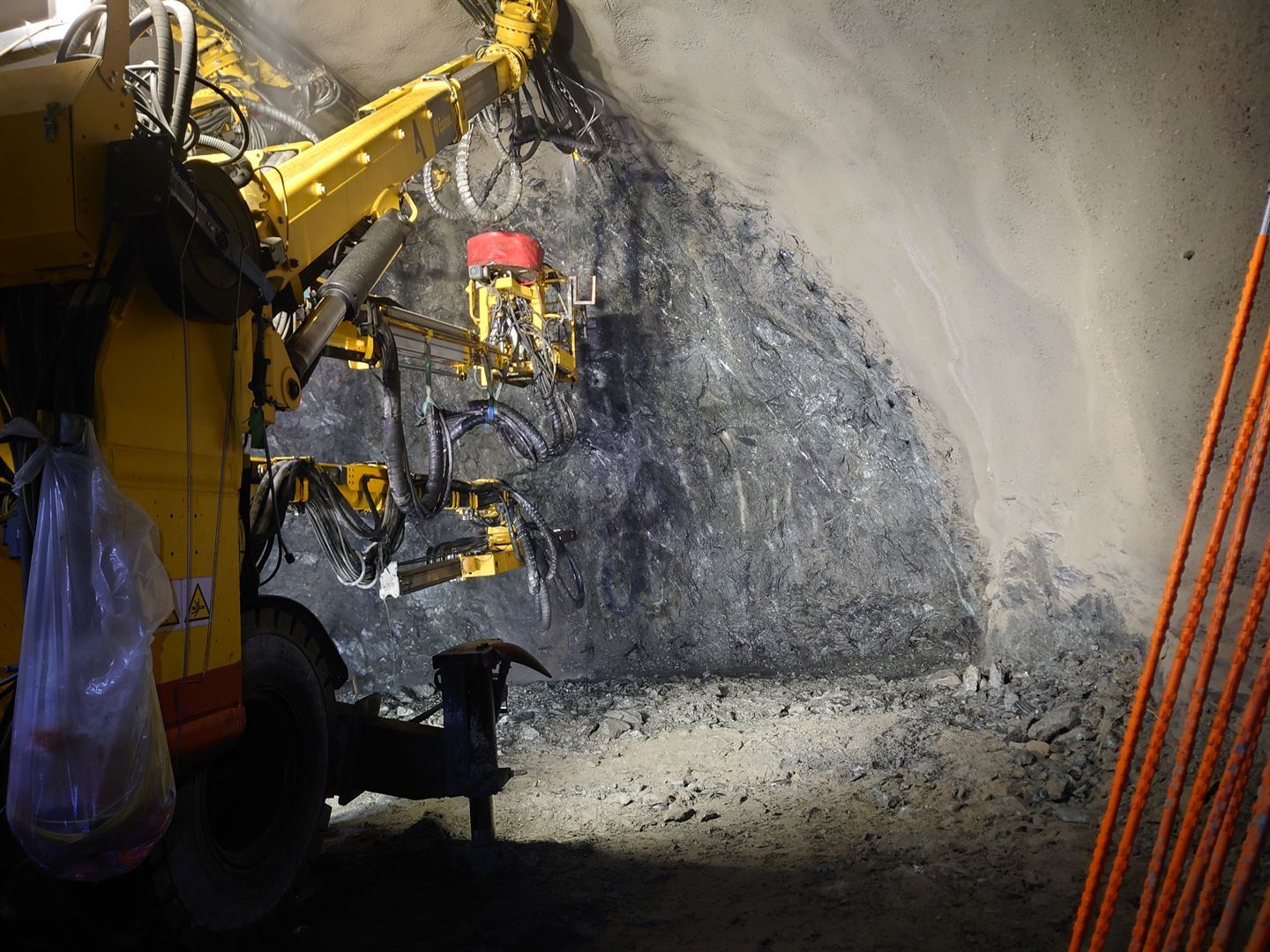 Gul boremaskin med fleire borarmar inne i ein tunnel, retta mot ein nyleg sprengt fjellvegg. Golvet er dekt av stein og grus, og tunneltaket er sikra med sprøytebetong.