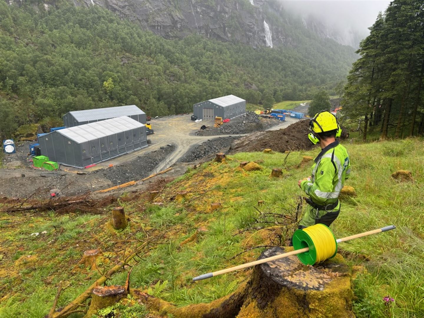 Ein anleggsarbeidar står i ei skogsli og ser ned på eit anleggsområde med riggbygg og maskiner i dalbotnen. Rundt området er det bratte fjellsider og tett skog.