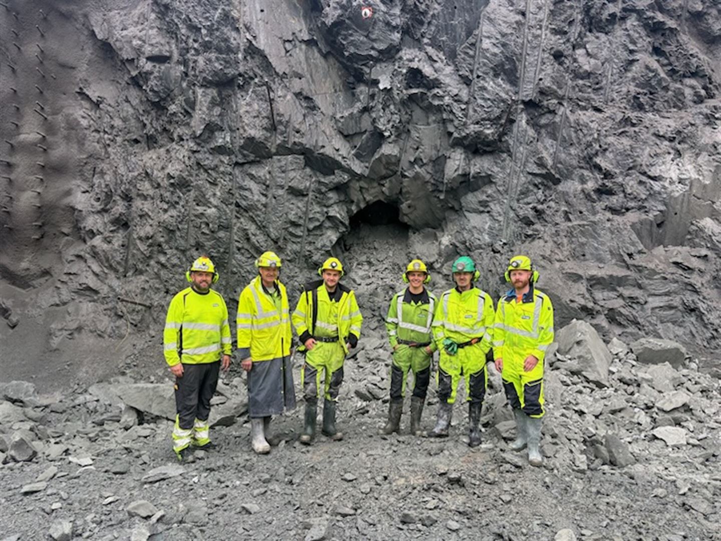 Seks anleggsarbeidarar i gule refleksklede og hjelm står framfor tunnelpåhogget inne i eit stein- og fjellanlegg.