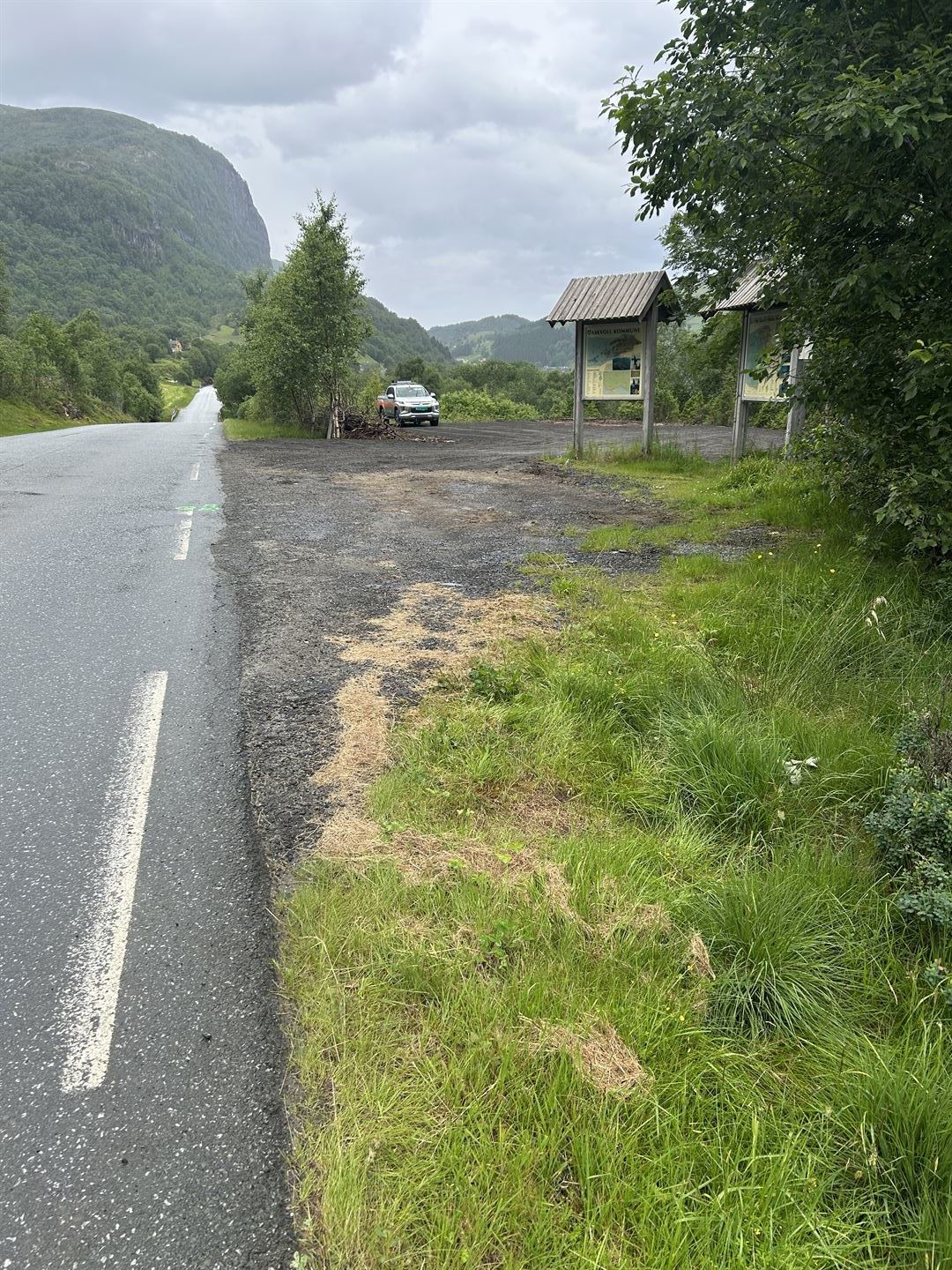 Nyleg opparbeidd grusplass i eit grønt fjellandskap. Skogkledde åsar og mørke regnskyer i bakgrunnen. Informasjonsskilt med kart står i forgrunnen.