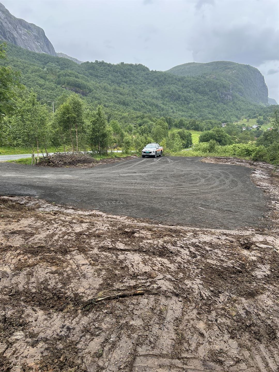 Nyleg opparbeidd grusplass i eit grønt fjellandskap. Ein bil står på plassen, med skogkledde åsar og mørke regnskyer i bakgrunnen.