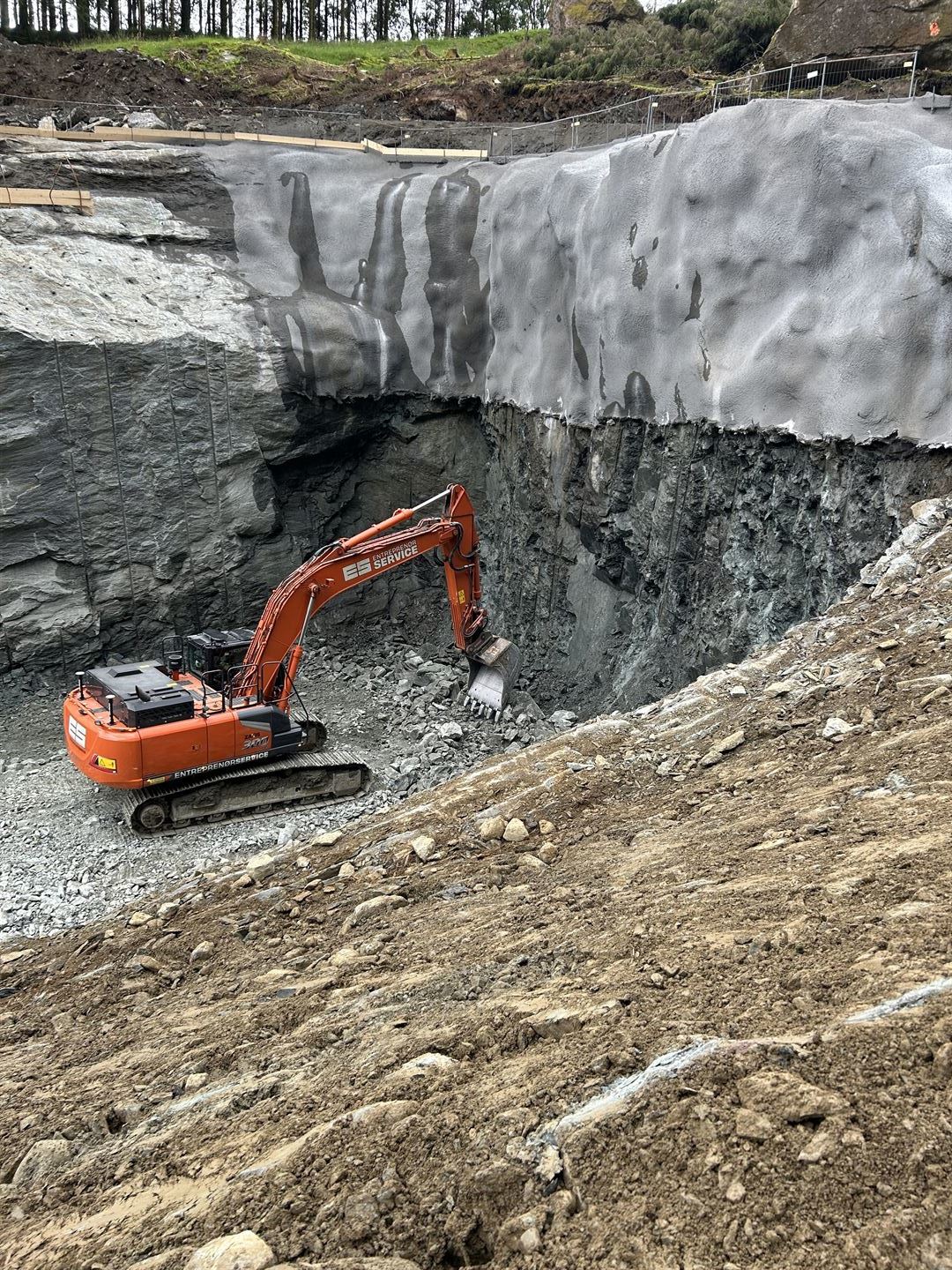 Gravemaskin arbeider i ei djup utsprengd byggegrop omgitt av bratte fjellveggar som er sikra med sprøytebetong.