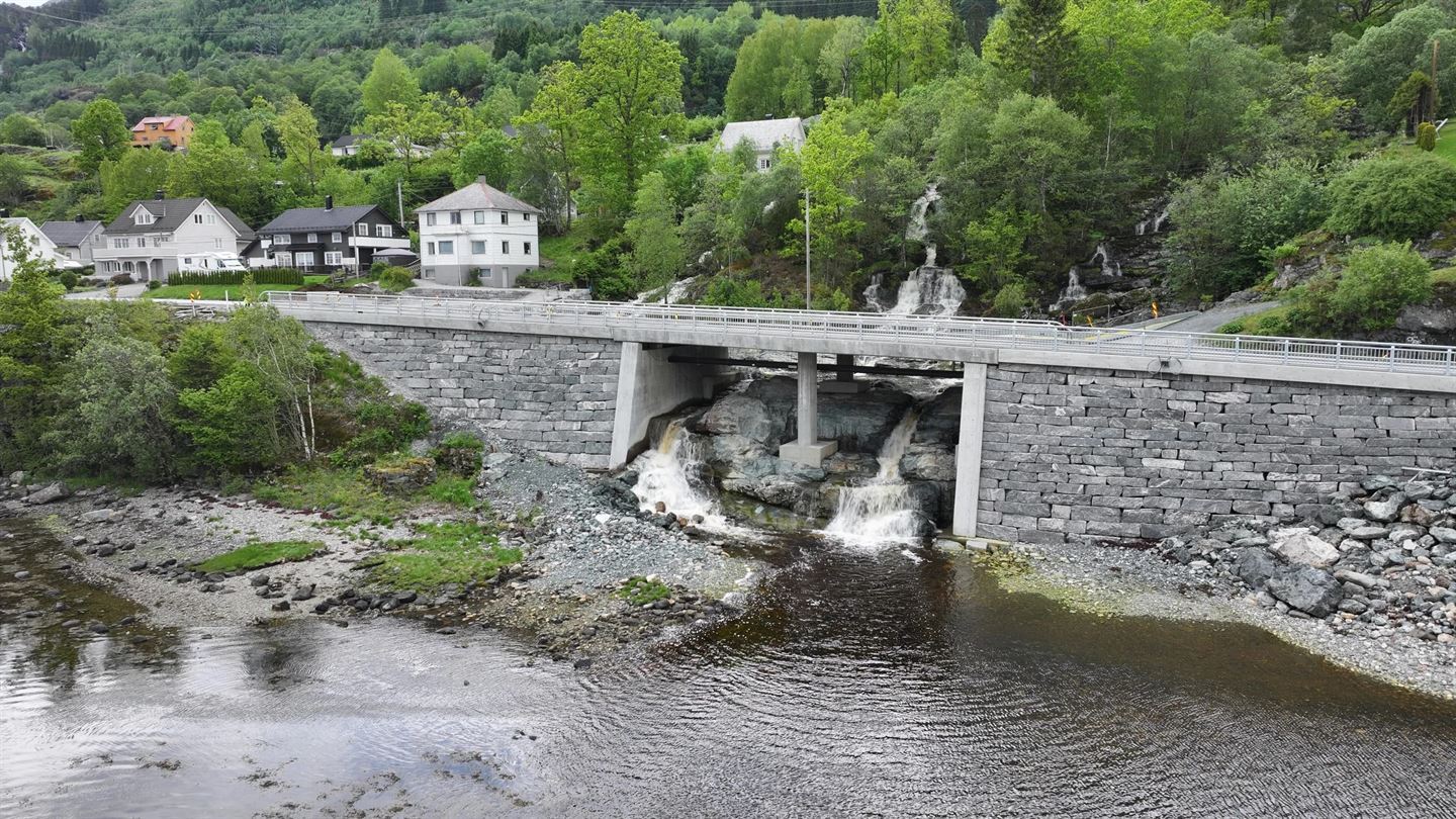 Dronebilete av ein ny betongbru med natursteinsmur på sidene. Under brua renn ein foss over nakne fjellhyllar og ut i ein mørk sjø. Grøne lauv­tre og bustadhus i bakgrunnen. Vegrekkverk i metall langs brukanten.