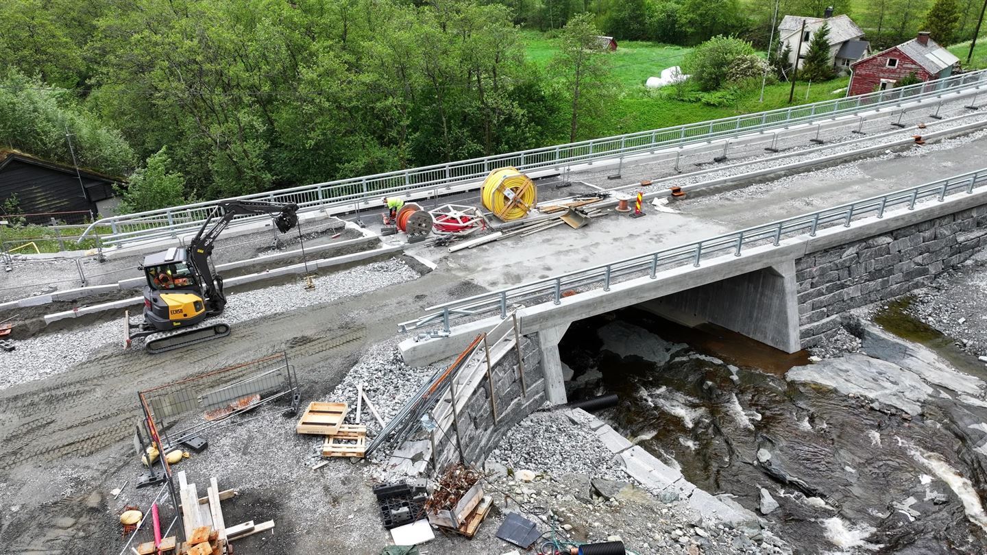 Dronebilete teke frå sida av ein betongbru som er under sluttføring. Ein gul gravemaskin står på brua, og det ligg kabeltromlar, stillas og byggjemateriell på bruplata. Under brua renn ei elv. Grøne tre og bygningar i bakgrunnen.