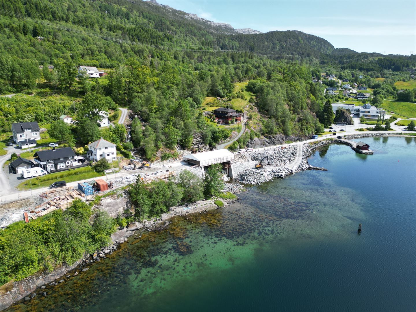 Naturskjønt kystområde med en blanding av bolighus og naturlig grøntområder. En vei går langs kysten, med flere hus plassert nær veien. Kystlinjen har steinete strender og klart vann, med noe vegetasjon synlig under vann. I bakgrunnen er det åser dekket av tett skog.