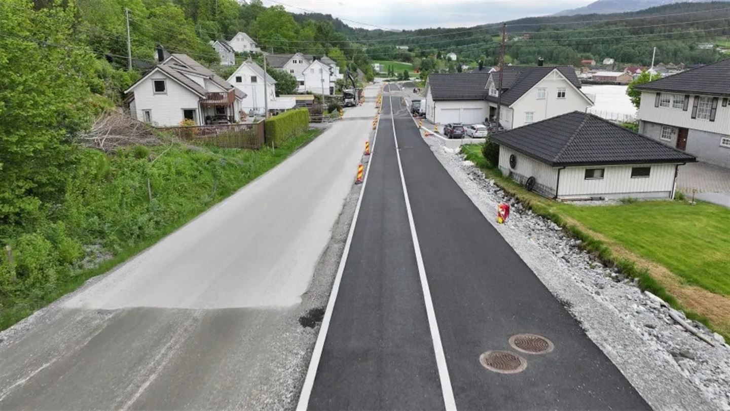 Dronebilete av pågåande asfalteringsarbeid langs ein veg med fjorden i bakgrunnen. Ein gul Volvo-maskin legg asfalt, og det er synleg gul miljøslange langs bergskjæringa til venstre. Arbeidsvarslingsskilt og anleggsutstyr langs vegen. Bustadhus og sjø i bakgrunnen.