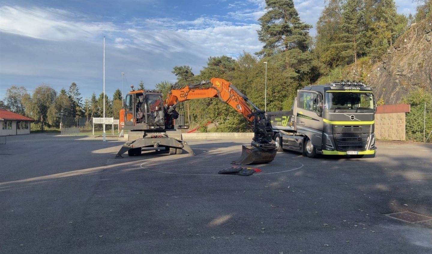 Ein svart lastgebil med gul stripe på står ved sida av ein oransje gravemaskin på ein skuleplass dekka med svart asfalt. 