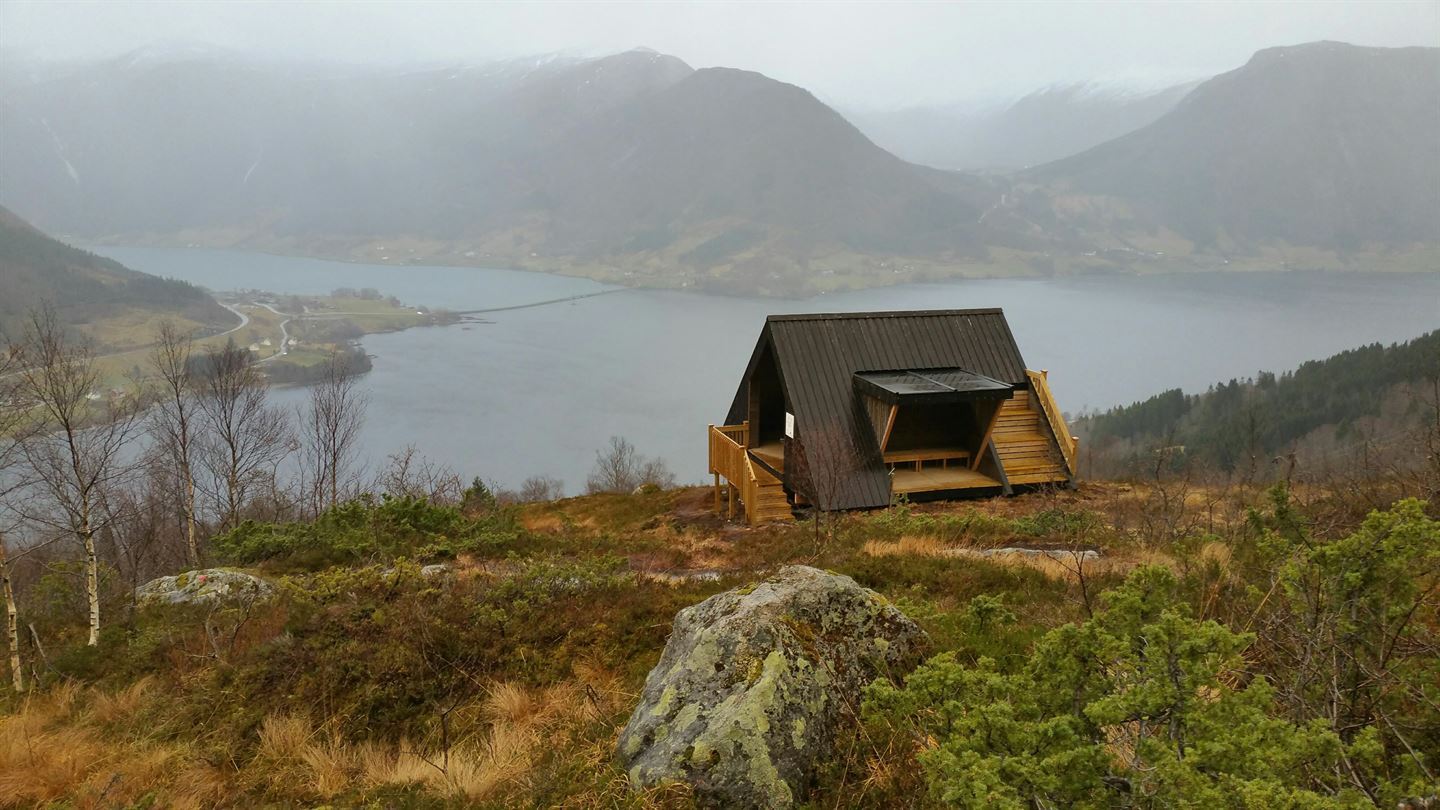 Bilde av ei dagsturhytte. I bakgrunnen er det Jølstravatnent og fjell. Regntung dag.