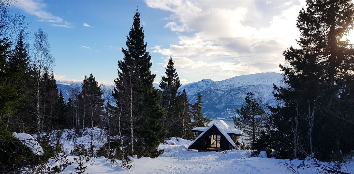 Bilde av ei dagsturhytte. Det er snø på taket og snø rundt hytta. Det er sol og litt skyer på himmelen. 