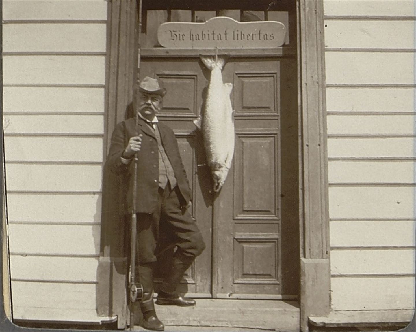 Svart-kvitt-bilde av mann med bart, hatt og fiskestong. Ved sidan av han heng ein stor fisk på ei dør.