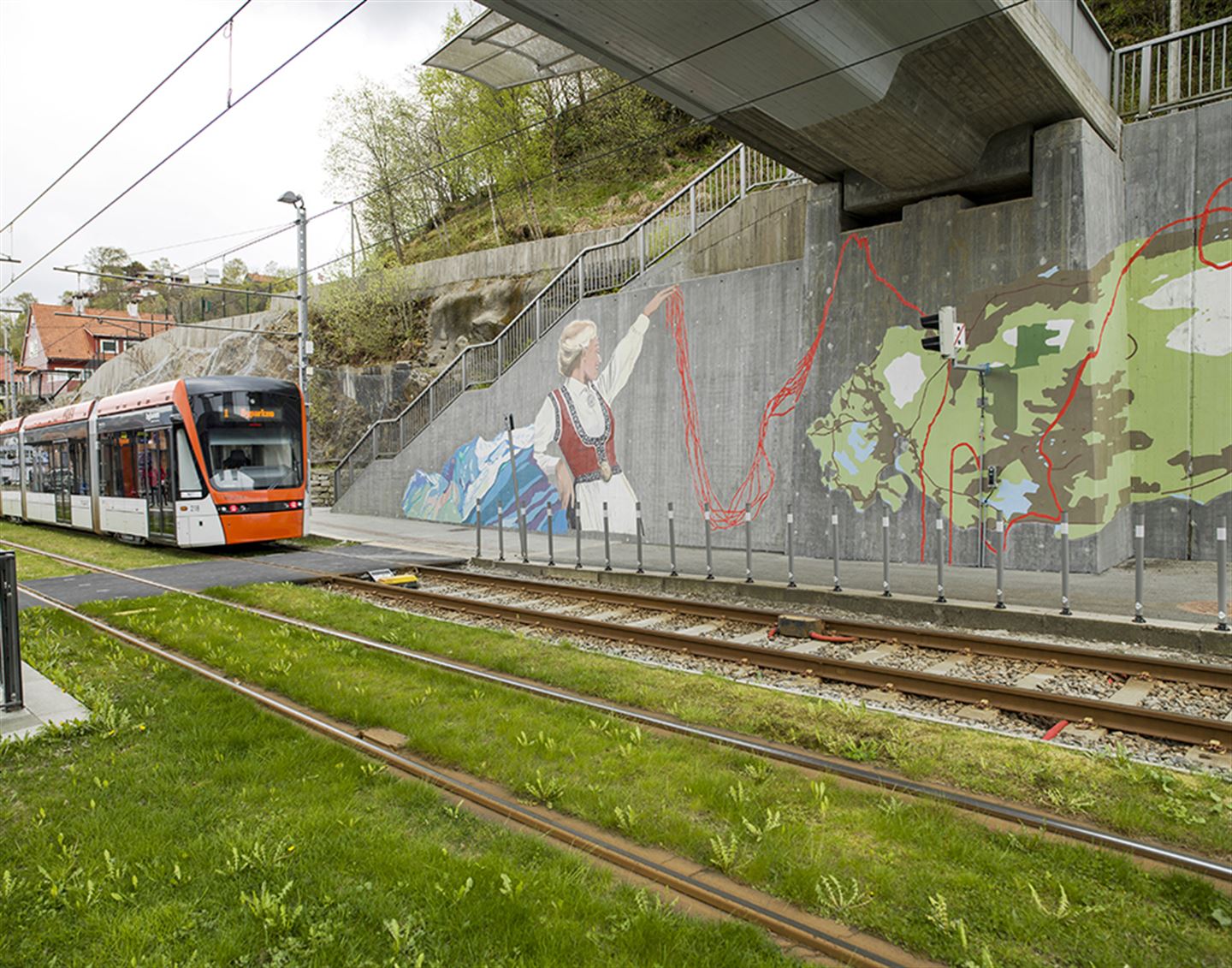 Foto av bybanen og kunstverket 1: 1 000 483 langs bybanetraseen. Kunstverket er målt på betong og syner ei kvinne i bunad, eit blått felt, eit grønt felt og raude strekar.