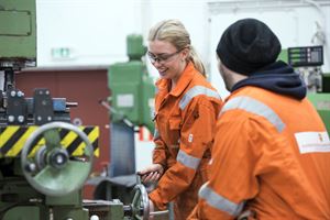 To elevar ved Rubbestadnes vidaregåande skule jobbar i verkstad ikledd oransje arbeidsklede. Kvinneleg elev har langt lyst hår, vernebriller og smiler medan ho arbeider med industrimaskin, mannleg elev ser på. 