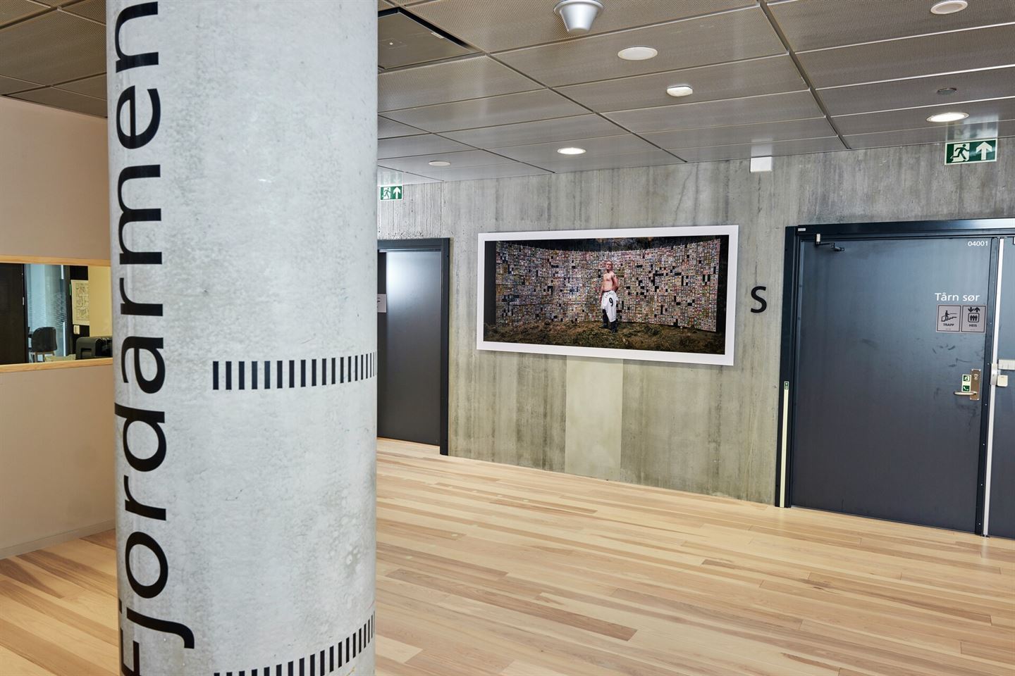 Stort fotografi av mann i fotballdrakt på vegg i kontorlandskap med smårom. I framgrunnen står ei betongsøyle med teksten Fjordarmen.
