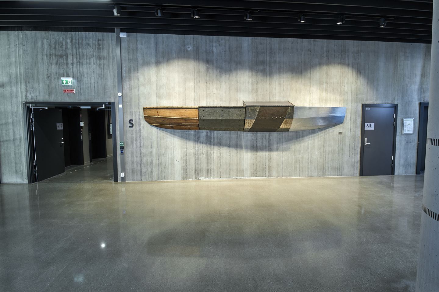 Foto av skulptur av halv båt på betongvegg. Båten er laga av tre, skiferstein og rustfritt stål.