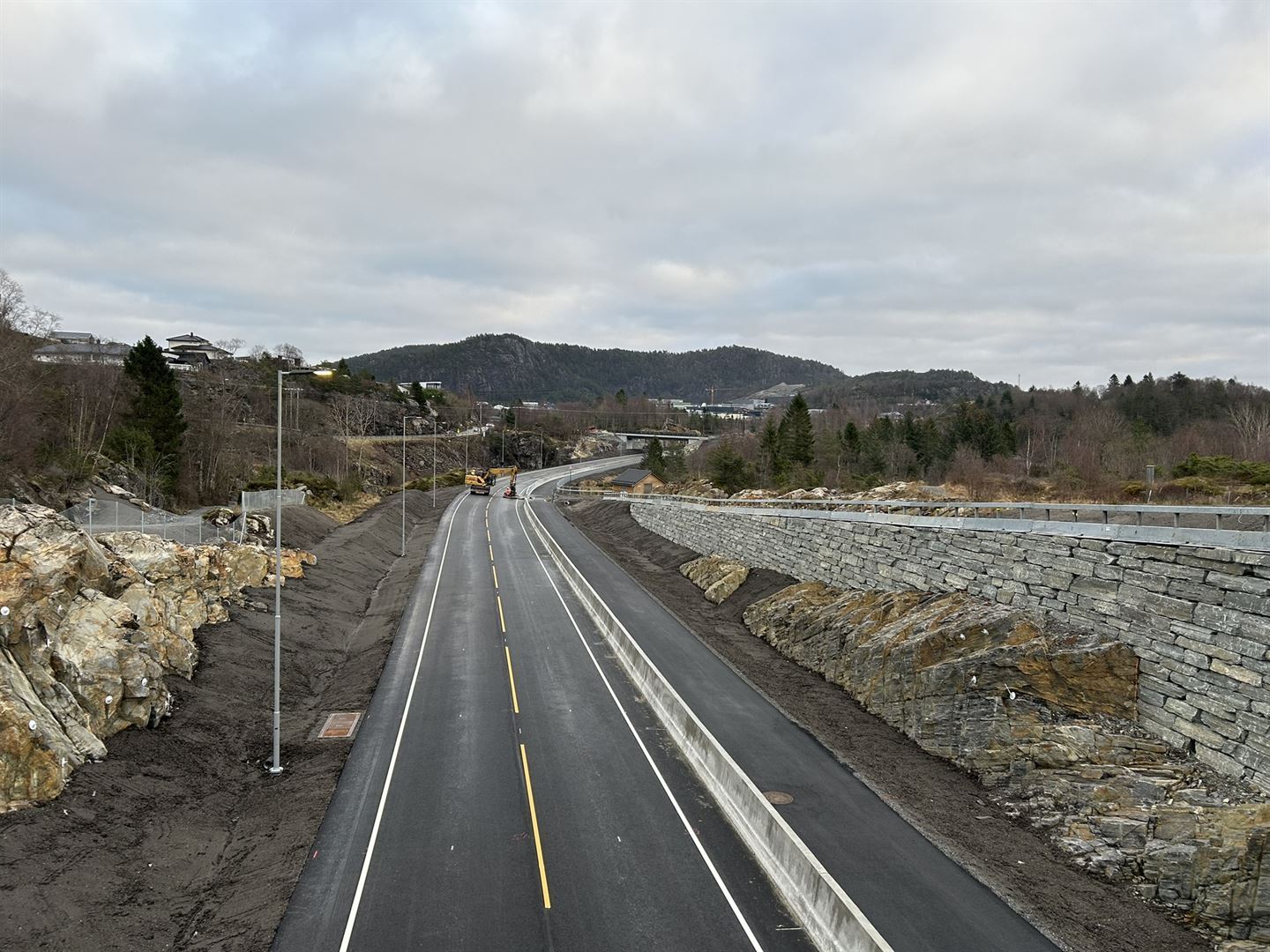 Ferdig veg med ny asfalt og gul stripe som går gjennom landskapet. Ein tørrmur på høgre side, og ein gravemaskin som står på asfalten midt i vegen.