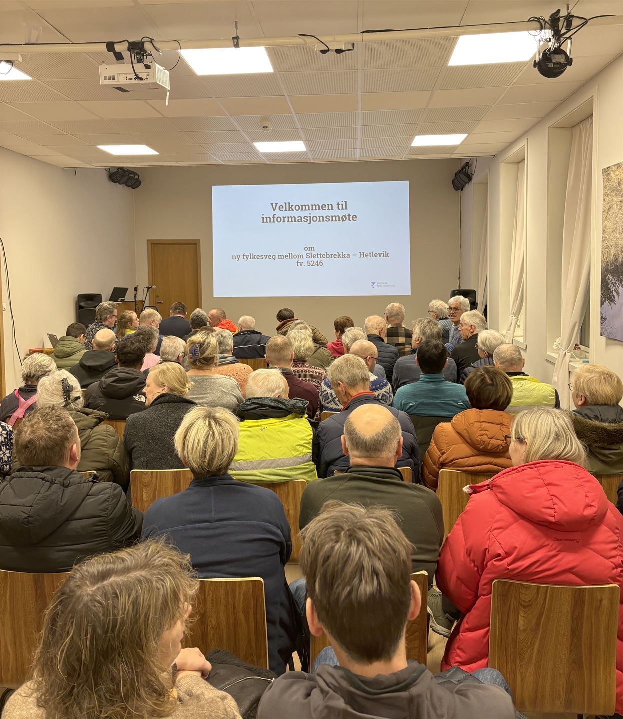 Samling av folk i en sal. På veggen heng det ein presentasjon.