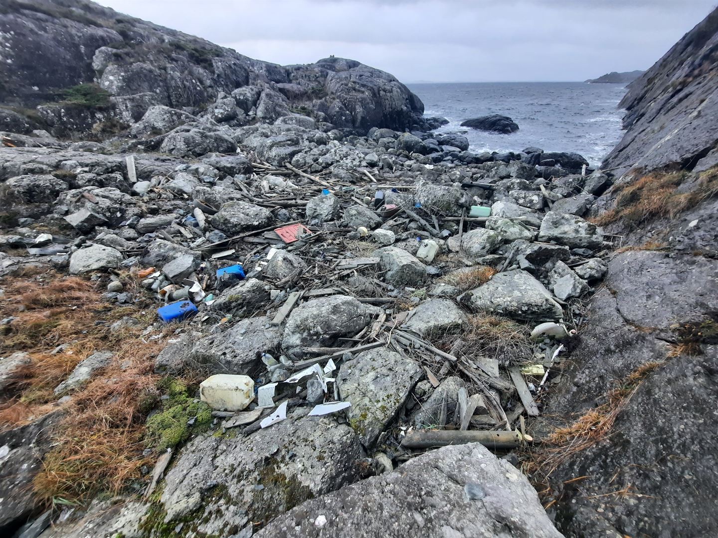 Strandlinje inne i ei vik, med havet rett utanfor. Store mengder plastsøppel, garn og andregjenstander ligg mellom steinane på stranda.