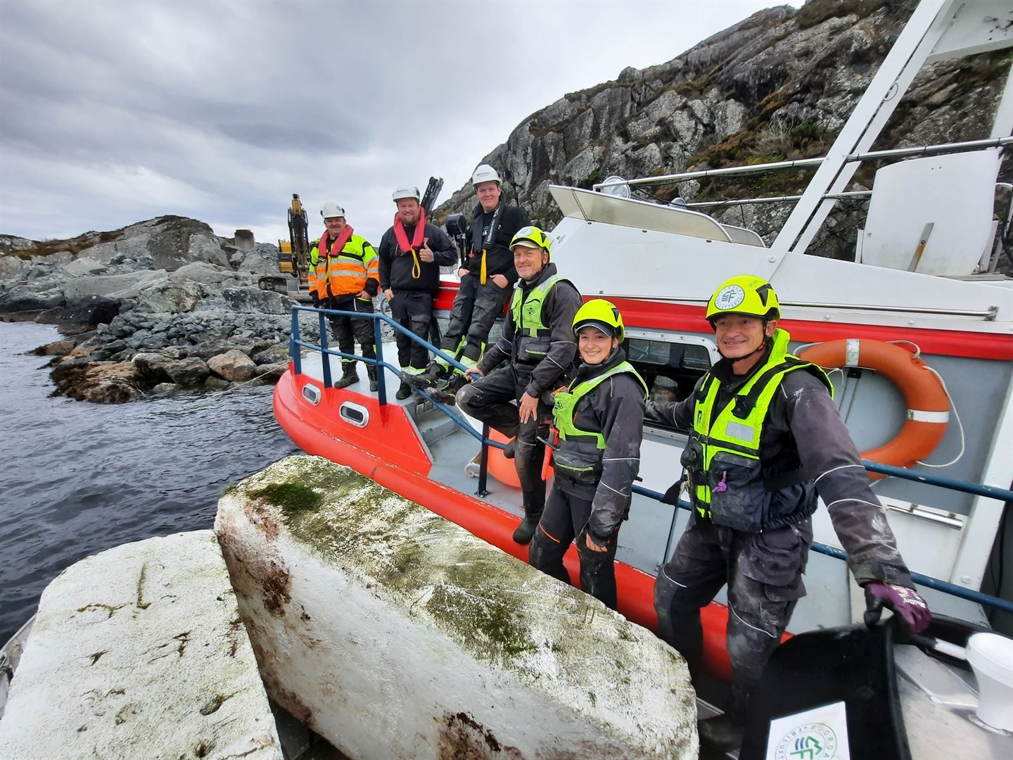 Seks smilande menneske som står lang rekka på ein båt som ligg ved kai. Alle har flytesetar og hjelmar på hovudet.