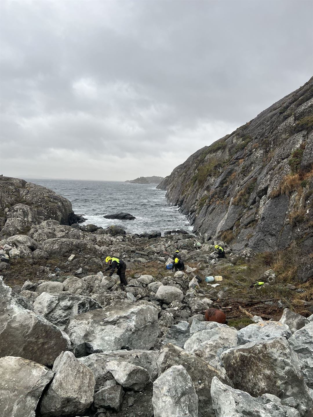 Ei steinstrand i ei vik, med havet rett utanfor. Fire menneske med gule hjelmar og gule vestar plukkar søppek som ligg blant steinane på stranda.