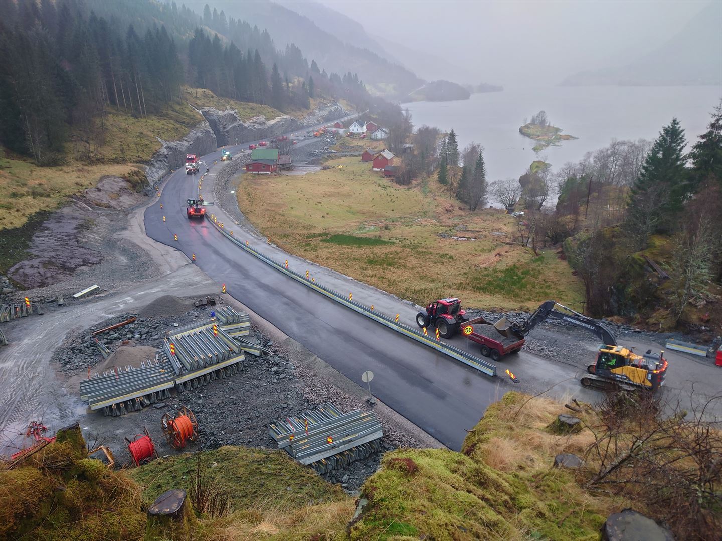 Veg langs fjord, med svart, nylagd asfalt. Mange anleggsmaskiner som jobbar på vegen. Mellom vegen og fjorden er det gule bøar, nokre hus og løer.
